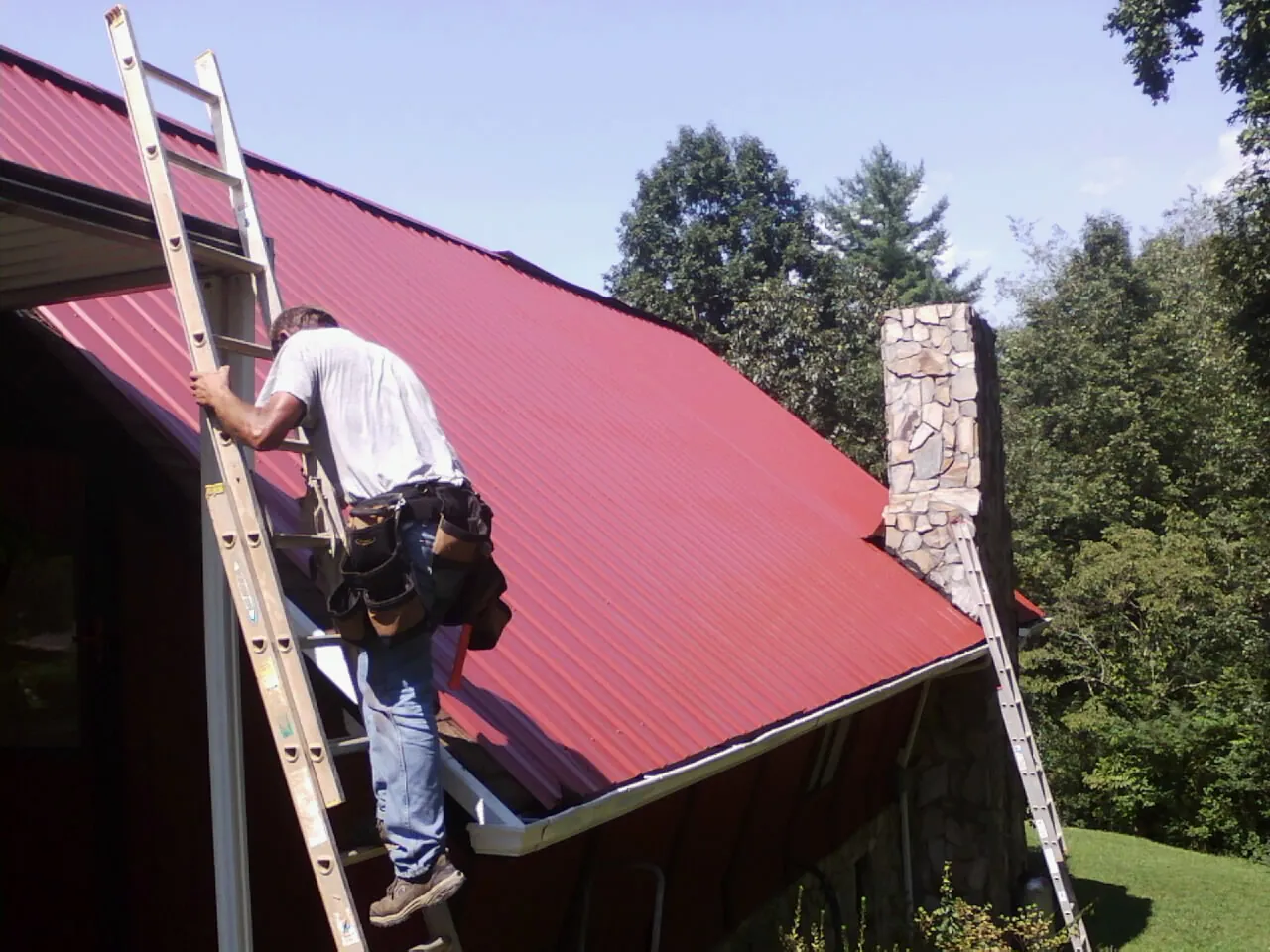 Roofer on a ladder beside a red metal roof and stone chimney — Soffit & Fascia Repair by Roofing Brothers in Griffith
